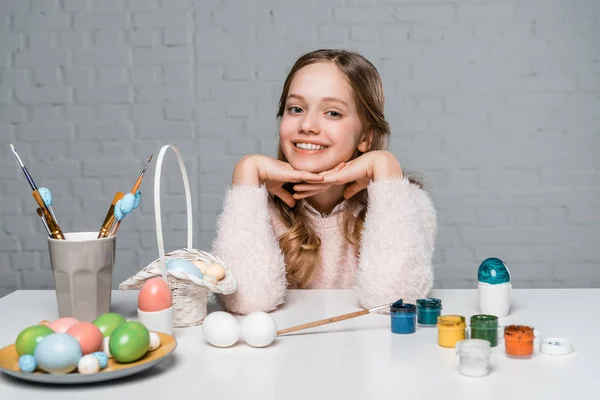 Чарівна щаслива дівчина посміхається на камеру, сидячи за столом з фарбами, кошиком і великодніми яйцями — стокове фото