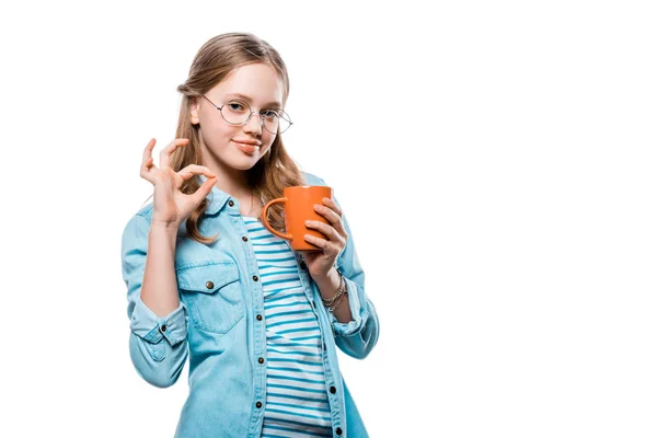 Beautiful girl in eyeglasses holding mug of tea and showing ok sign isolated on white — Stock Photo