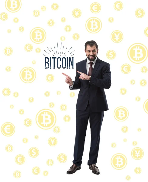 Barbudo sonriente hombre de negocios en traje apuntando a la palabra bitcoin aislado en blanco - foto de stock