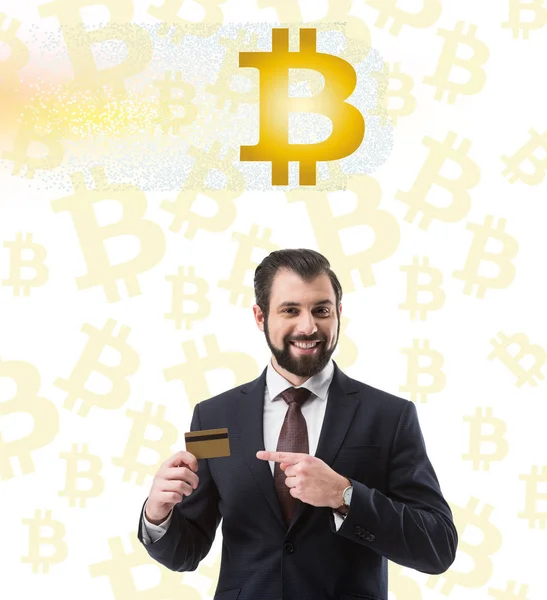 Sonriente hombre de negocios apuntando a la tarjeta de crédito, símbolos bitcoin en blanco - foto de stock