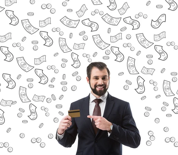 Sonriente hombre de negocios apuntando a la tarjeta de crédito, billetes de dólar y monedas símbolos aislados en blanco — Stock Photo