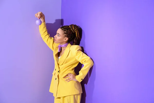 Chica afroamericana posando en traje amarillo con manzana púrpura, sobre fondo ultra violeta de moda - foto de stock