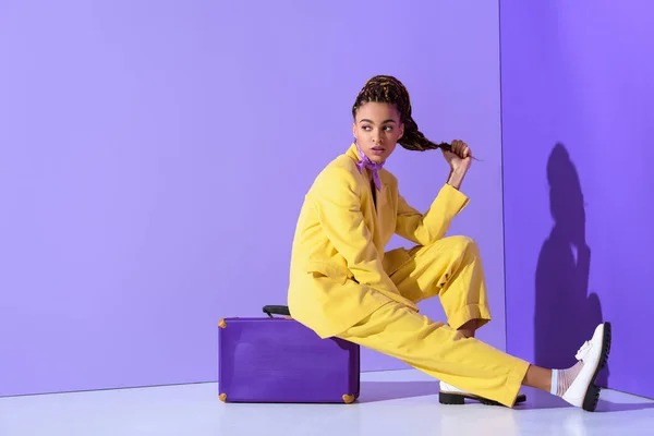 African american girl posing in yellow suit sitting on purple suitcase, on trendy ultra violet background — Stock Photo