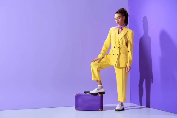 Attrayant afro-américain fille posant en costume jaune avec valise violette, sur fond ultra violet à la mode — Photo de stock