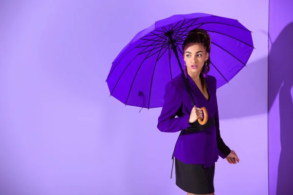 Chica afroamericana elegante posando con paraguas púrpura en la pared ultravioleta - foto de stock