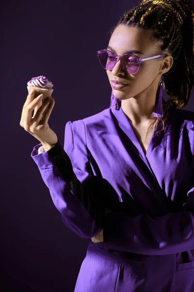 Стильная африканская американка в ультрафиолетовой куртке и солнцезащитных очках, смотрящая на кексы, изолированные на темно-фиолетовый — стоковое фото