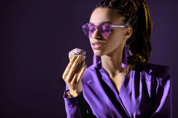 Stylish african american girl in ultra violet jacket and sunglasses with cupcake, isolated on dark purple — Stock Photo
