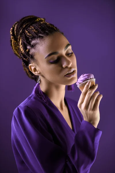 Sensual menina americana africana em casaco roxo olhando para cupcake, isolado em roxo — Fotografia de Stock