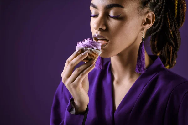 Apasionado afroamericano chica en chaqueta púrpura comer cupcake, aislado en púrpura - foto de stock