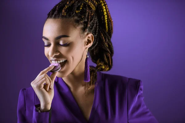 Cheerful mulatto girl eating macaron, isolated on purple — Stock Photo