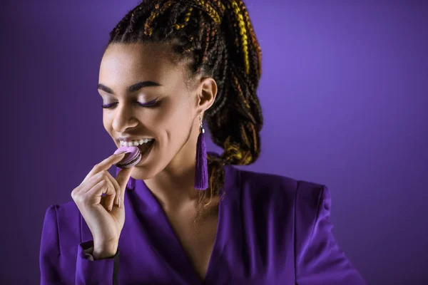 Menina mulata alegre comer macaron, isolado em roxo — Fotografia de Stock