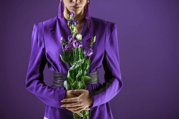 Обрезанный вид модной девушки, позирующей в костюме с букетом цветов, изолированной на ультрафиолетовом — стоковое фото