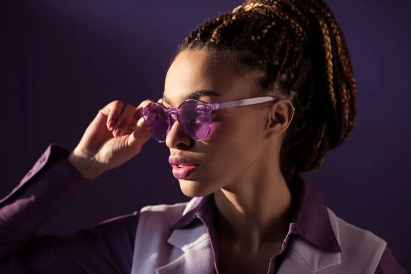 Elegante chica afroamericana posando en gafas de sol púrpura, aislado en púrpura - foto de stock