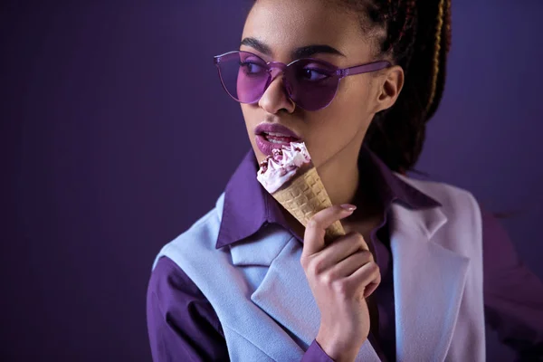 Африканская американская девушка позирует в фиолетовых солнцезащитных очках с мороженым в конусе, изолированный на фиолетовый — стоковое фото