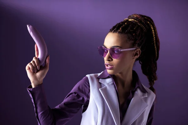 Menina americana africana elegante bonita em óculos de sol roxos segurando banana como arma, isolado em roxo — Fotografia de Stock