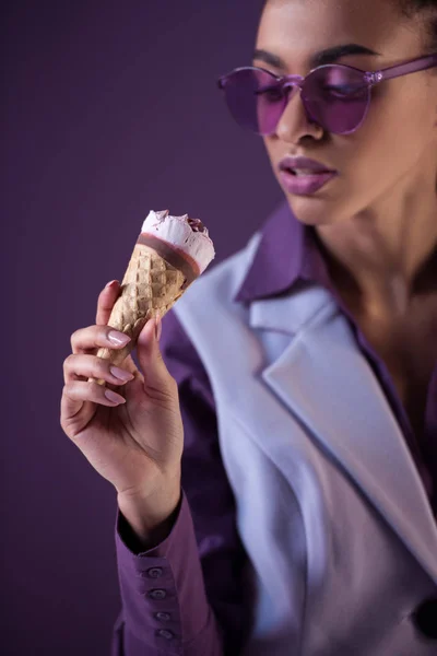 Belle fille afro-américaine en lunettes de soleil ultra violettes à la mode regardant la crème glacée en cône, isolée sur violet — Photo de stock