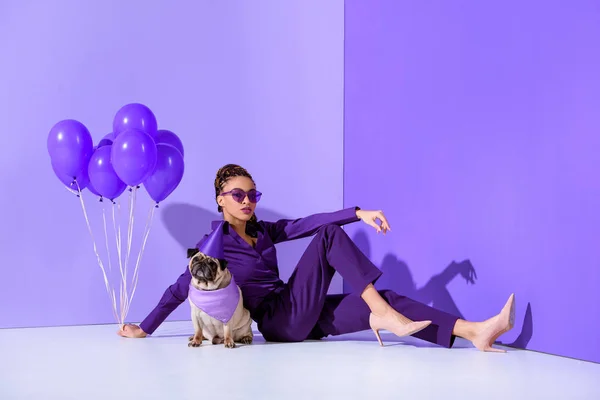 Elegante chica afroamericana posando con globos púrpura y pug, tendencia ultra violeta — Stock Photo