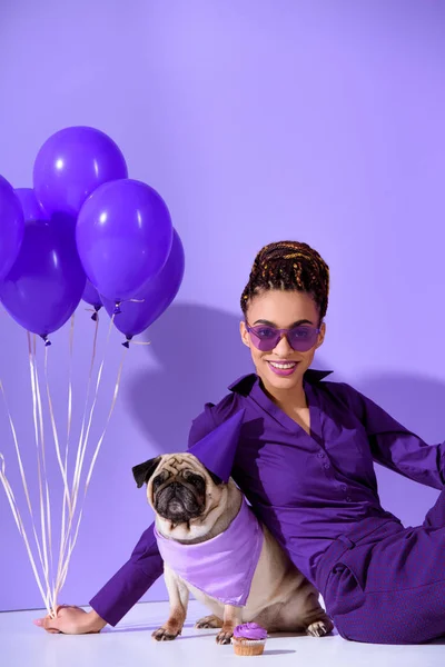 Smiling african american girl posing with purple balloons and pug, ultra violet trend — Stock Photo