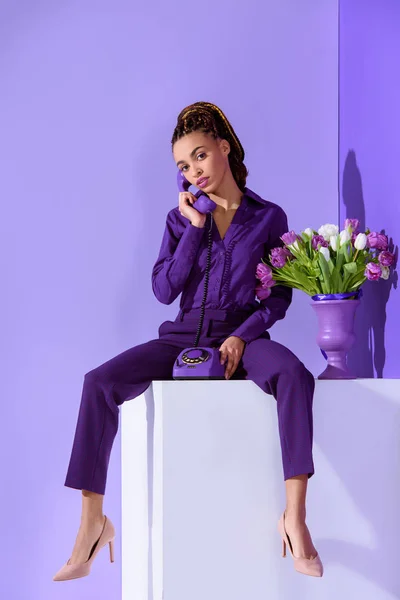 Elegante chica mulata hablando por teléfono rotatorio y sentado en cubo con ramo de tulipanes - foto de stock