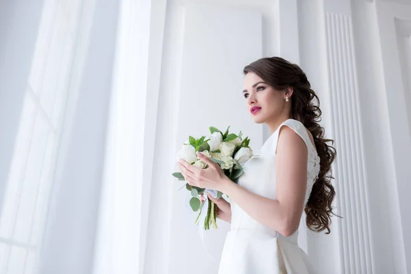 Красивая молодая невеста со свадебным букетом, смотрящая в окно — стоковое фото