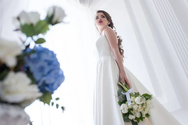 Vista inferior de la novia posando en vestido blanco con ramo de boda - foto de stock