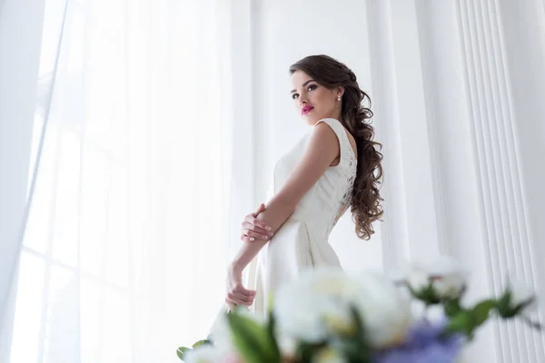 Brunette caucasian bride in wedding dress at window, flowers on foreground — Stock Photo