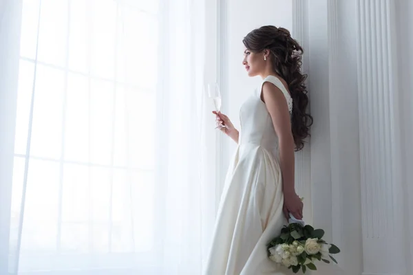 Brunette bride in dress with wedding bouquet and glass of champagne looking at window — Stock Photo