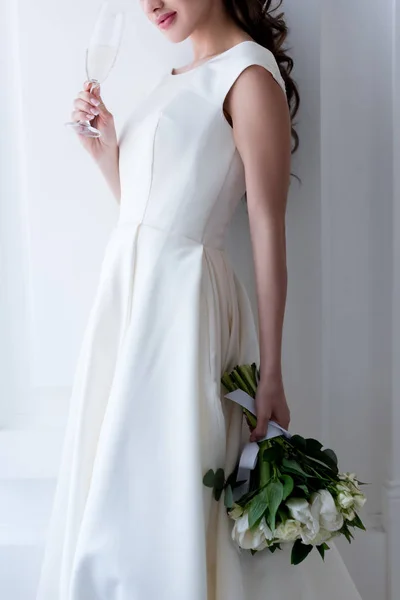 Cropped view of bride in elegant dress with wedding bouquet and glass of champagne — Stock Photo