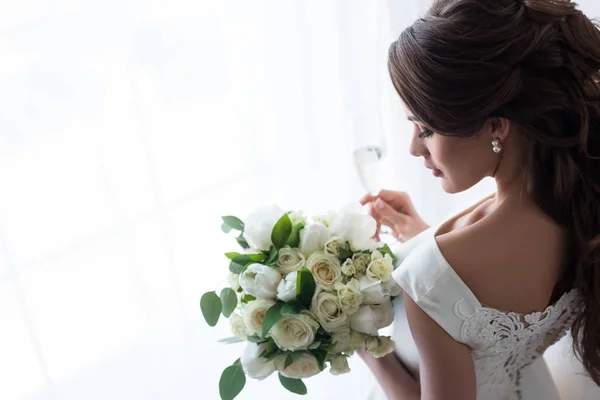 Attractive bride in traditional dress with wedding bouquet and glass of champagne standing at window — Stock Photo