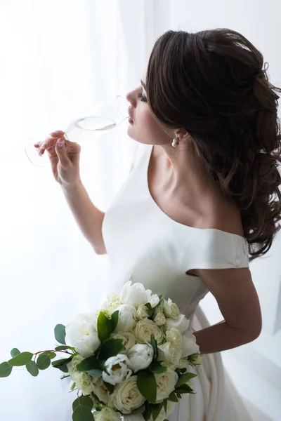 Hermosa joven novia con ramo de boda beber champán - foto de stock