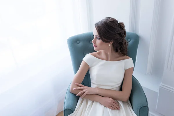 Morena mujer en vestido de novia sentado en sillón y mirando a la ventana - foto de stock