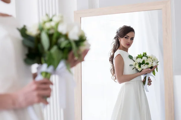Beautiful bride in dress with wedding bouquet looking at her reflection in mirror — Stock Photo