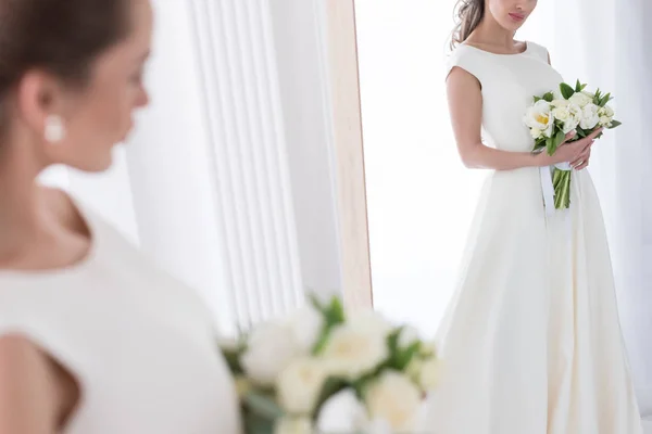 Bride in traditional dress with wedding bouquet looking at her reflection in mirror — Stock Photo