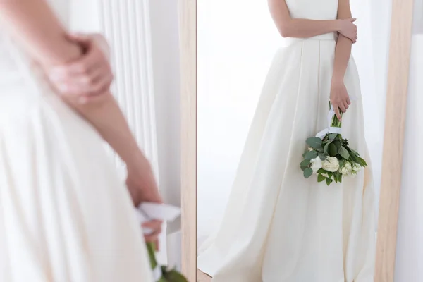 Vista recortada de la novia en vestido tradicional con ramo de boda mirando su reflejo en el espejo - foto de stock