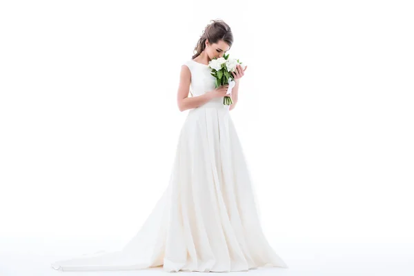 Элегантная невеста позирует в традиционном платье и нюхает свадебный букет, изолированный на белом — стоковое фото