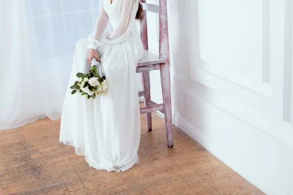 Cropped view of elegant bride sitting on big chair with wedding bouquet — Stock Photo