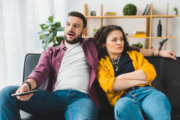 Angry woman lying on boyfriend shoulder while he using remote controller — Stock Photo