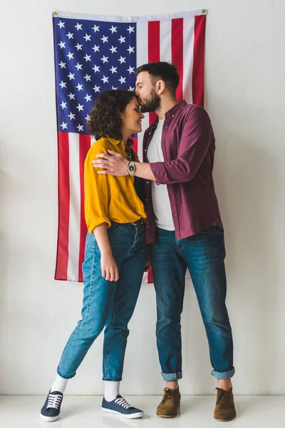 Uomo baciare la fronte fidanzata davanti al muro con bandiera americana — Foto stock