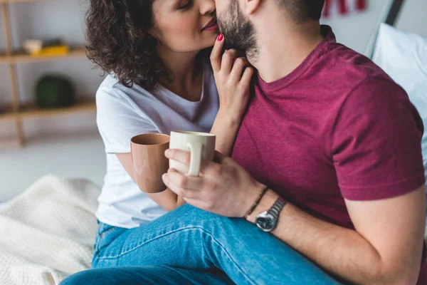 Recortado vista de sonriente mujer besando novio mientras él sosteniendo tazas - foto de stock