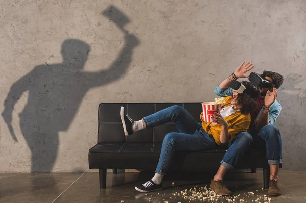Sombra de hombre sosteniendo hacha y pareja asustada con palomitas de maíz usando auriculares de realidad virtual - foto de stock