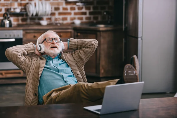 Hombre viejo feliz en los auriculares usando el ordenador portátil con los pies en la mesa - foto de stock