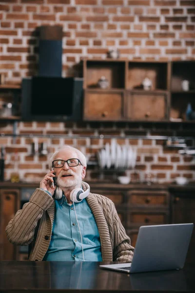 Hombre mayor en auriculares hablando por teléfono por ordenador portátil - foto de stock