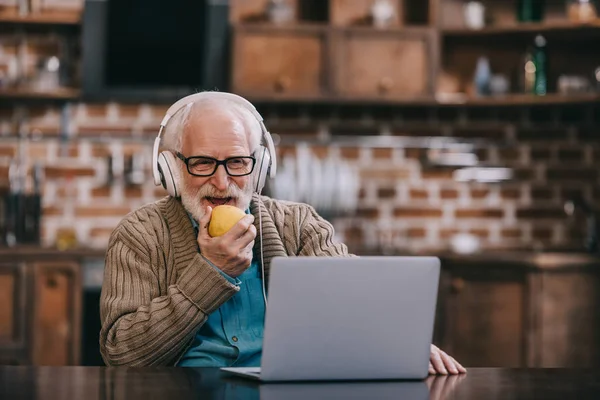 Hombre mayor feliz en los auriculares comiendo manzana usando el ordenador portátil - foto de stock