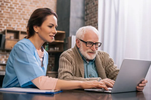 Enfermera sentada por el hombre mayor usando el ordenador portátil - foto de stock