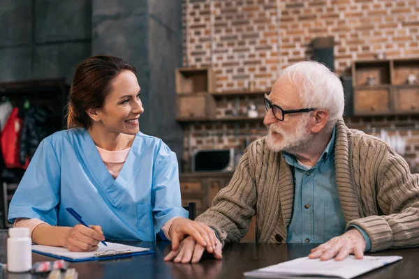 Nurse writing down senior patient medical complaints and holding his hand — Stock Photo