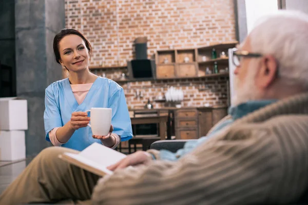 Infirmière donnant tasse avec boisson au patient âgé avec livre — Photo de stock