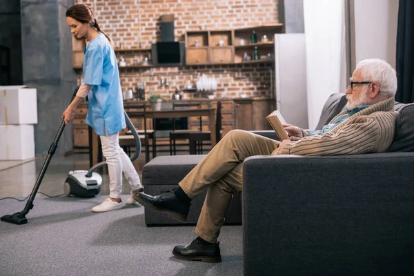 Senior man reading while caregiver vacuums room — Stock Photo