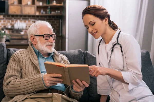 Doctor and senior man reading book on sofa — Stock Photo