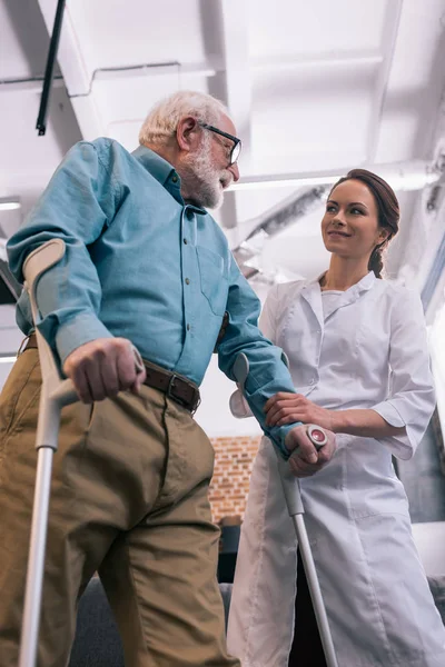 Senior man leaning on crutches and smiling female doctor — Stock Photo