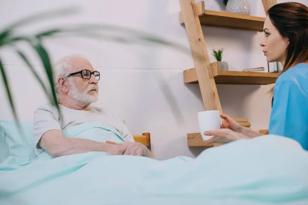 Nurse giving cup with drink to senior patient in bed — Stock Photo